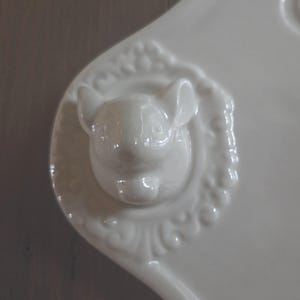 May include: A white ceramic serving dish with a raised, detailed bunny head in the center. The bunny is surrounded by a decorative, embossed border. The dish has a smooth, glossy finish and a curved handle.