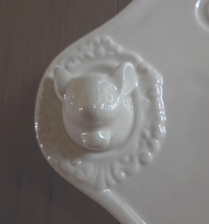 May include: A white ceramic serving dish with a raised, detailed bunny head in the center. The bunny is surrounded by a decorative, embossed border. The dish has a smooth, glossy finish and a curved handle.