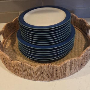 May include: A round woven seagrass tray with scalloped edges and integrated handles, containing two stacked sets of circular plates. The plates feature a cream-colored center with a wide, dark blue rim and a thin, light blue inner border.