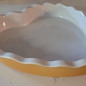 Op de afbeelding: Een hartvormige keramische ovenschaal met een geschulpte rand. De schaal heeft een lichtgele buitenkant en een witte binnenkant. De rand is wit. Geschikt om in te bakken en te serveren.