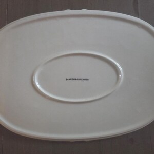 May include: Off-white oval serving platter with a scalloped edge. The platter has a raised oval center and the text "by Anthropologie" printed in black. The platter is made of ceramic.