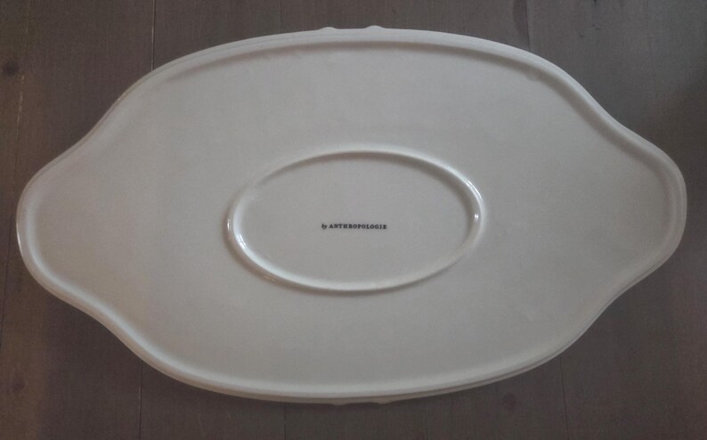 May include: Off-white oval serving platter with a scalloped edge. The platter has a raised oval center and the text "by Anthropologie" printed in black. The platter is made of ceramic.