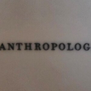 May include: The phrase "by ANTHROPOLOGIE" is displayed in a dark gray, stylized font. The text is centered on a light gray background.
