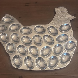 May include: A silver-colored metal egg tray shaped like a chicken. The tray features twenty-one oval indentations for holding eggs, with a detailed embossed design of a chicken head and comb at the top right corner of the platter.