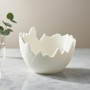 Op de afbeelding: Een witte, eivormige decoratieve kom met een unieke, gekartelde rand. De kom staat op een marmeren oppervlak, met een kleine vaas met groen in de achtergrond. Het ontwerp van de kom voegt een moderne touch toe aan de woondecoratie.