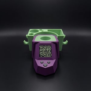 Puede incluir: Un dispositivo morado y verde impreso en 3D con el texto "DAB RITE PRO" en la parte delantera. El dispositivo tiene forma rectangular con bordes redondeados y tres botones debajo de la pantalla. Está conectado a una bandeja verde con múltiples compartimentos.