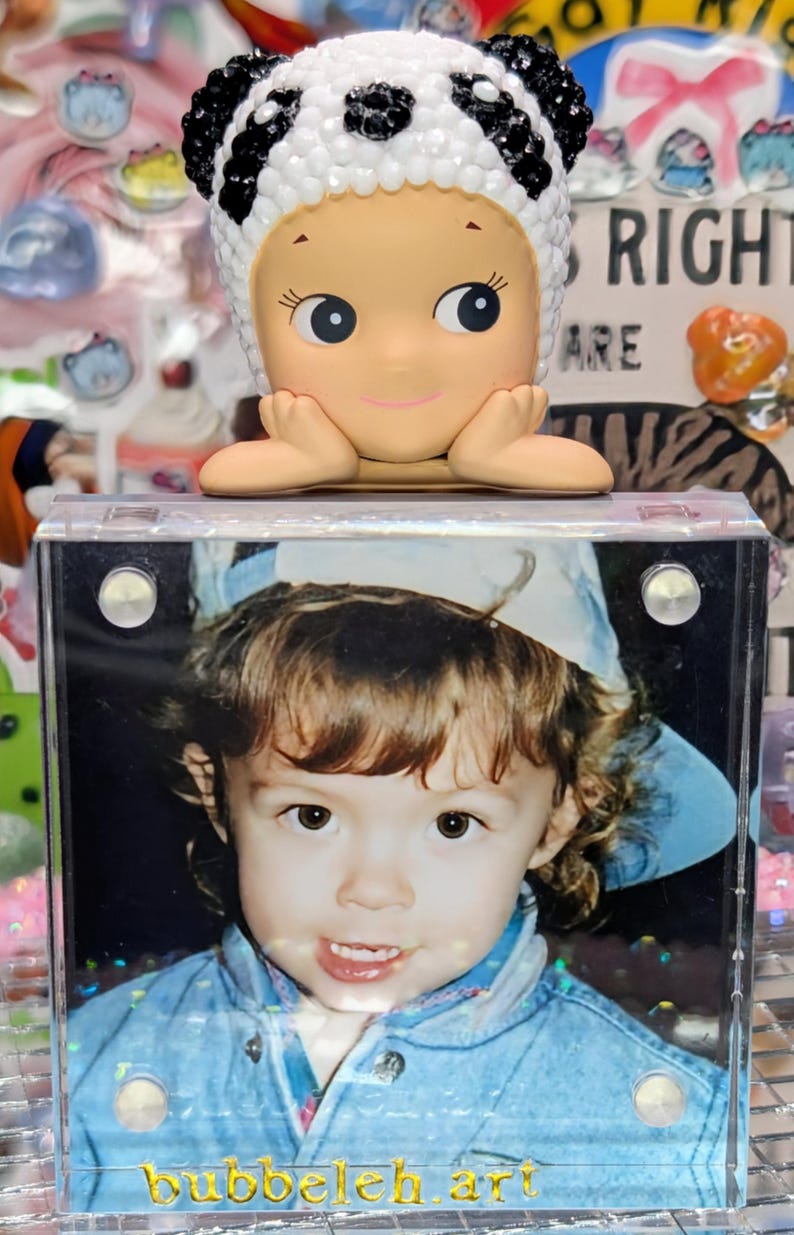 May include: A figurine of a baby-like character wearing a panda hat, resting on a clear acrylic frame. The frame holds a photograph of a young child in a denim jacket. The text "bubbeleh.art" is visible on the frame.