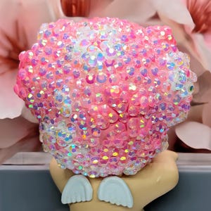 May include: A pink decorative object covered in iridescent rhinestones. The object has a rounded shape and is adorned with numerous sparkling crystals. The base of the object is a light tan color with two white, wing-shaped accents.