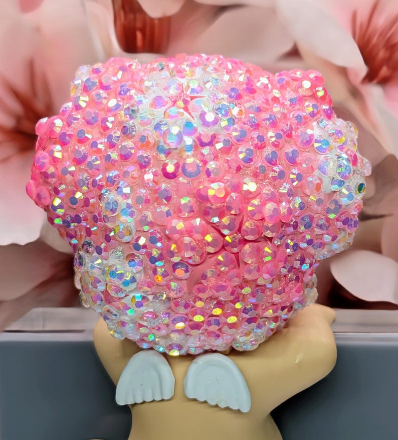May include: A pink decorative object covered in iridescent rhinestones. The object has a rounded shape and is adorned with numerous sparkling crystals. The base of the object is a light tan color with two white, wing-shaped accents.