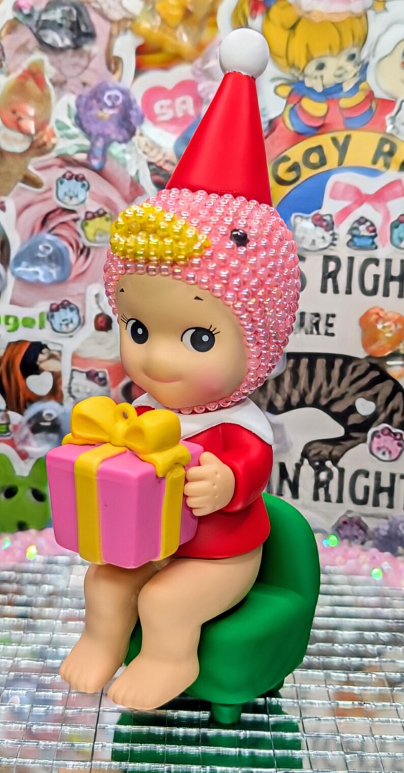 May include: A seated figurine wearing a red outfit and a pink beaded cap with a white pom-pom. The figurine holds a pink gift box with a yellow bow. It sits on a green chair. The background is covered in stickers.
