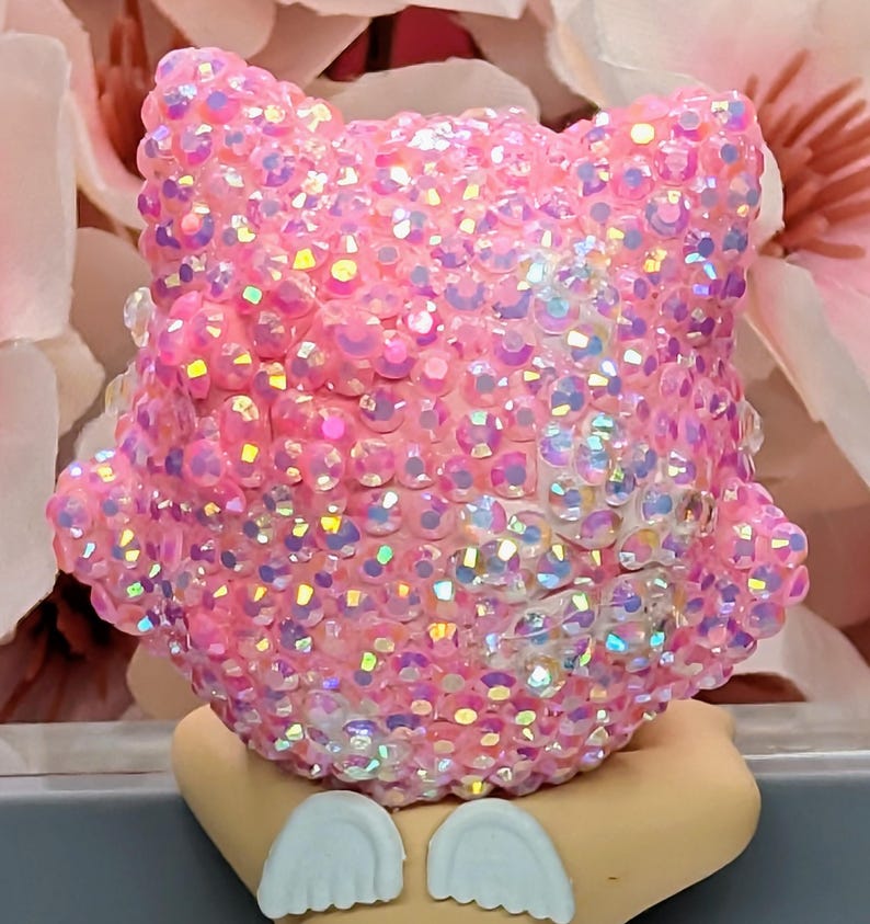 K&ouml;nnte beinhalten: Eine rosa, mit Strass besetzte Eulenfigur mit schillernden Strasssteinen. Die Eule hat gro&szlig;e, runde Augen und einen abgerundeten K&ouml;rper. Die Figur steht auf einem hellbraunen Sockel mit wei&szlig;en fl&uuml;gelf&ouml;rmigen Details.