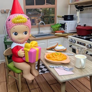 May include: A figurine of a character wearing a red and white outfit and a pink and yellow hat, holding a pink and yellow gift box. The scene is set in a miniature kitchen with a table set for a meal, including pasta, toast, and a mug.