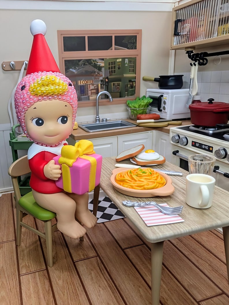 May include: A figurine of a character wearing a red and white outfit and a pink and yellow hat, holding a pink and yellow gift box. The scene is set in a miniature kitchen with a table set for a meal, including pasta, toast, and a mug.