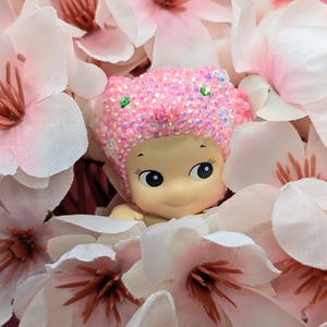 May include: A small, smiling doll with a pink, sequined cap peeks out from a bed of light pink and white artificial flowers. The doll has large, expressive eyes and a cheerful expression, creating a whimsical and charming scene.