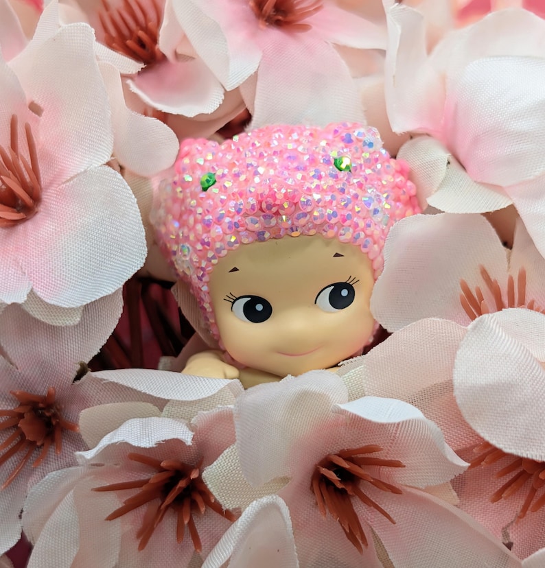 May include: A small, smiling doll with a pink, sequined cap peeks out from a bed of light pink and white artificial flowers. The doll has large, expressive eyes and a cheerful expression, creating a whimsical and charming scene.