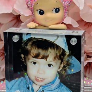 May include: A decorative figurine with a pink glittery head and a smiling face rests atop a clear acrylic block. The block displays a photo of a child in a blue jacket, with the text "bubbeleh.art" at the bottom.