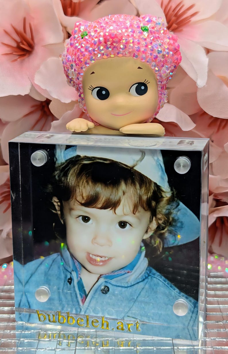 May include: A decorative figurine with a pink glittery head and a smiling face rests atop a clear acrylic block. The block displays a photo of a child in a blue jacket, with the text "bubbeleh.art" at the bottom.
