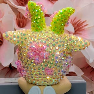 K&ouml;nnte beinhalten: Eine skurrile Figur, die mit schillernden Pailletten bedeckt ist. Die Figur hat gelbe, rosa und gr&uuml;ne Akzente, mit gr&uuml;nen, paillettenbesetzten, hornartigen Vorspr&uuml;ngen. Die Figur ruht auf einem hellen Sockel mit wei&szlig;en fl&uuml;gelf&ouml;rmigen Details.