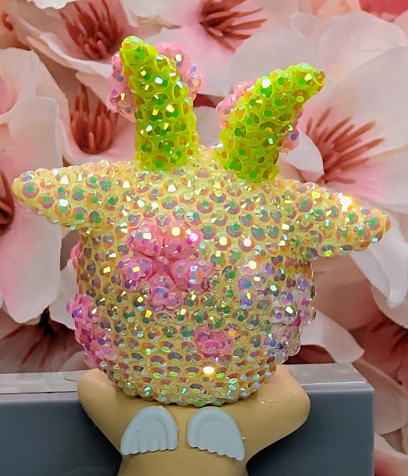 K&ouml;nnte beinhalten: Eine skurrile Figur, die mit schillernden Pailletten bedeckt ist. Die Figur hat gelbe, rosa und gr&uuml;ne Akzente, mit gr&uuml;nen, paillettenbesetzten, hornartigen Vorspr&uuml;ngen. Die Figur ruht auf einem hellen Sockel mit wei&szlig;en fl&uuml;gelf&ouml;rmigen Details.