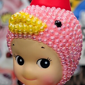 May include: A close-up of a doll with a pink beaded cap and a red cone-shaped hat. The doll has large, round eyes and a smiling expression. Yellow beads accent the cap. The doll is a collectible item.