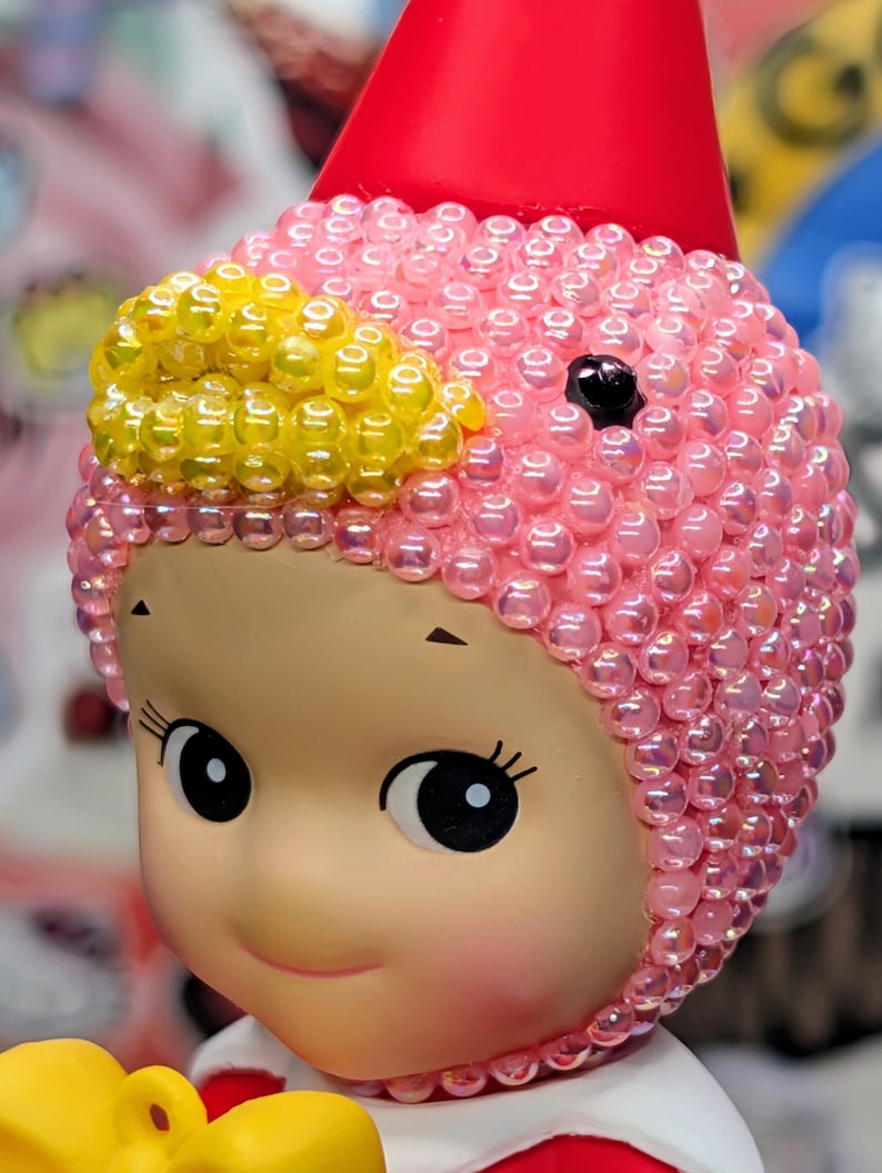 May include: A close-up of a doll with a pink beaded cap and a red cone-shaped hat. The doll has large, round eyes and a smiling expression. Yellow beads accent the cap. The doll is a collectible item.