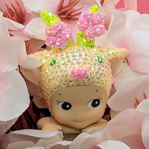 K&ouml;nnte beinhalten: Figur einer l&auml;chelnden, baby&auml;hnlichen Figur mit gelbem, schillerndem Kopfschmuck und rosa Blumenakzenten. Die Figur ist von rosa und wei&szlig;en Kunstblumen umgeben, die einen weichen, floralen Hintergrund bilden.