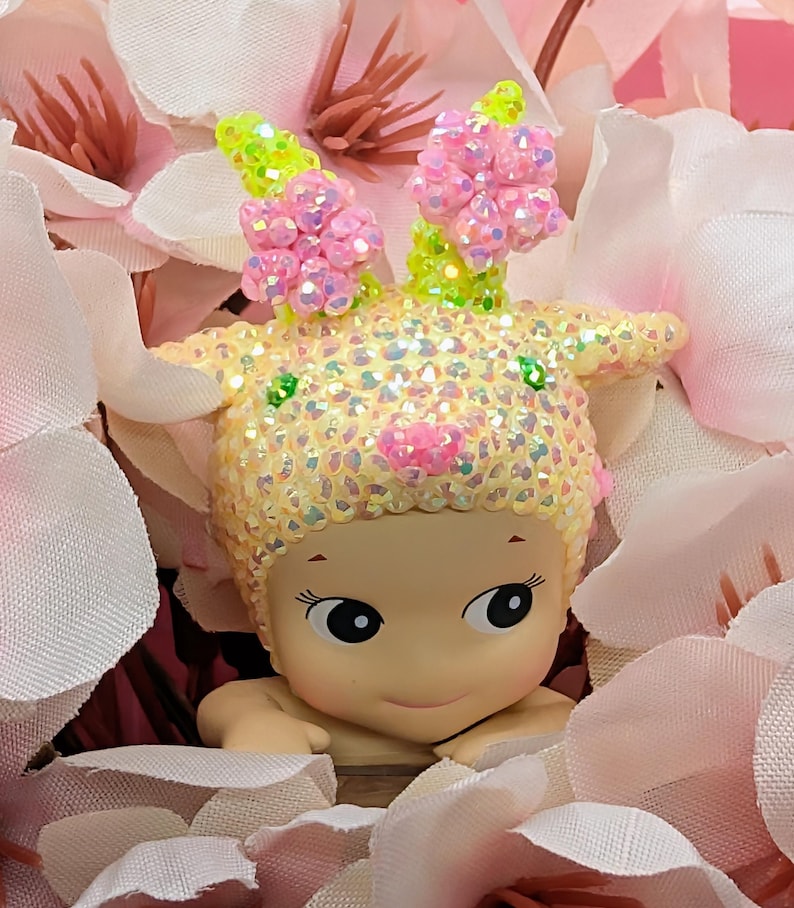 K&ouml;nnte beinhalten: Figur einer l&auml;chelnden, baby&auml;hnlichen Figur mit gelbem, schillerndem Kopfschmuck und rosa Blumenakzenten. Die Figur ist von rosa und wei&szlig;en Kunstblumen umgeben, die einen weichen, floralen Hintergrund bilden.