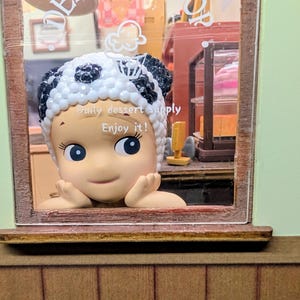May include: A miniature dessert shop scene featuring a smiling figure wearing a panda-themed hat. The figure is framed by a window, with the text "Daily dessert supply. Enjoy it!" visible. The shop interior includes shelves and a sign that says "Dessert Shop".