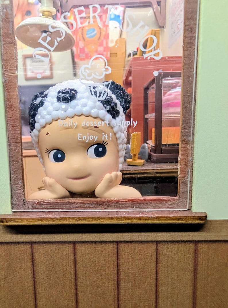May include: A miniature dessert shop scene featuring a smiling figure wearing a panda-themed hat. The figure is framed by a window, with the text "Daily dessert supply. Enjoy it!" visible. The shop interior includes shelves and a sign that says "Dessert Shop".