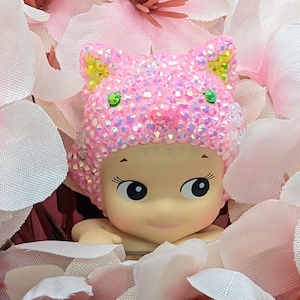 K&ouml;nnte beinhalten: Eine kleine Figur mit heller Haut, gro&szlig;en Augen und einem sanften L&auml;cheln, die einen rosa Katzenohrhut mit schillernden Strasssteinen tr&auml;gt. Die Figur ist von weichen rosa und wei&szlig;en Bl&uuml;tenbl&auml;ttern umgeben, was eine zarte und skurrile Szene schafft.