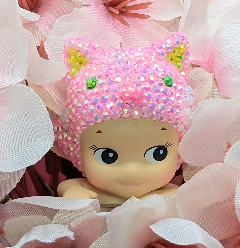 K&ouml;nnte beinhalten: Eine kleine Figur mit heller Haut, gro&szlig;en Augen und einem sanften L&auml;cheln, die einen rosa Katzenohrhut mit schillernden Strasssteinen tr&auml;gt. Die Figur ist von weichen rosa und wei&szlig;en Bl&uuml;tenbl&auml;ttern umgeben, was eine zarte und skurrile Szene schafft.