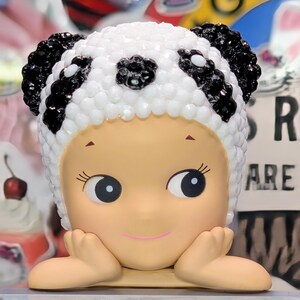May include: A figurine of a child-like figure with a panda bear hat. The hat is white with black accents and black gem-like embellishments. The figure has a light skin tone, large eyes, and is resting its chin on its hands.