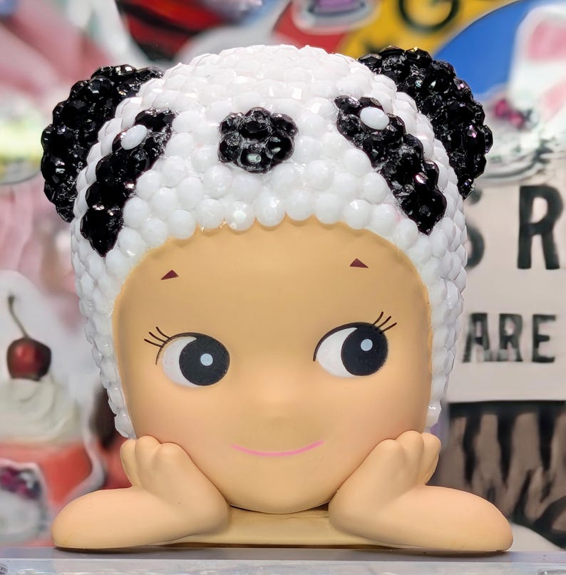 May include: A figurine of a child-like figure with a panda bear hat. The hat is white with black accents and black gem-like embellishments. The figure has a light skin tone, large eyes, and is resting its chin on its hands.
