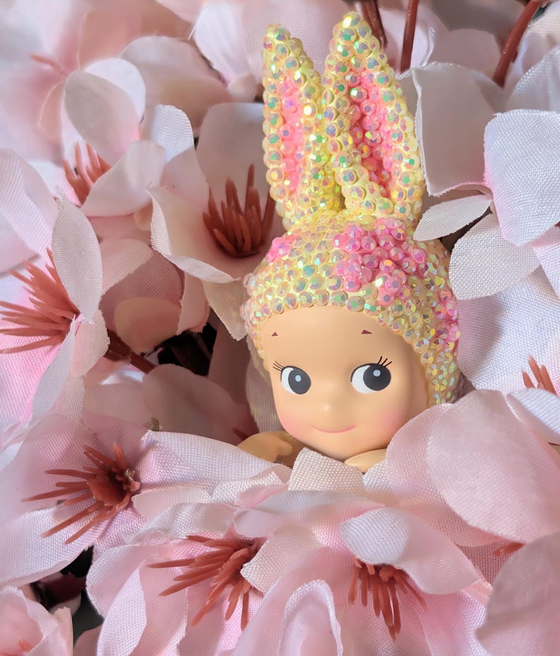 May include: A figurine of a baby with large eyes and a smiling expression, wearing a bunny ear headpiece. The headpiece and ears are adorned with iridescent sequins and pink accents. The figurine is nestled among soft pink and white artificial flowers.