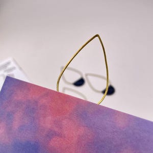 Può includere: Un segnalibro in metallo color oro a forma di goccia è inserito in una carta in stile acquerello viola e rosa. Lo sfondo presenta un paio di orecchini bianchi e blu navy.