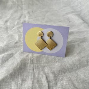 Könnte beinhalten: Ein Paar goldfarbene Ohrringe mit geometrischem Design. Jeder Ohrring hat einen kreisförmigen Aufsatz und einen rautenförmigen Anhänger mit radialen Linien. Die Ohrringe werden auf einer lavendelfarbenen Karte mit gelben und weißen Kreisen präsentiert.