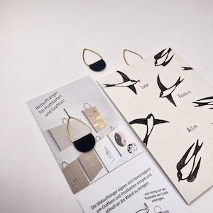 Può includere: Orecchini con montatura in metallo dorato e base semicircolare nera. Sono visibili anche due cartoline con illustrazioni di uccelli in bianco e nero e testo in tedesco. Una cartolina recita "Liebe", "Freiheit" e "Alles".