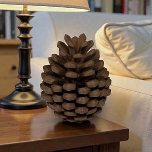May include: A brown, pine cone-shaped decorative object. The object is made of a series of overlapping, textured segments. A black band encircles the middle of the pine cone. The object is sitting on a wooden table next to a lamp.
