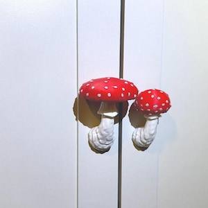 May include: Two mushroom-shaped cabinet pulls. The mushroom caps are red with white spots, and the stems are white. The pulls are mounted on a white cabinet door.