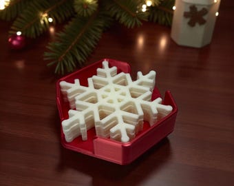 May include: A stack of white snowflake-shaped decorations in a red holder. The snowflakes are made of a light-colored material and are stacked neatly. The holder is a bright red color. Christmas tree and ornaments are in the background.
