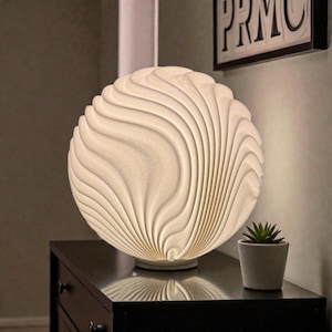 May include: A white, spherical lamp with a textured, wave-like design. The lamp is illuminated, casting a soft glow. It sits on a black nightstand next to a small potted succulent. A framed artwork is visible in the background.