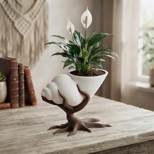 May include: A white ceramic planter shaped like a conch shell, supported by a brown, root-like base. The planter holds a green peace lily plant with two white spathe flowers. The structure is designed to hold soil and a plant securely in place.