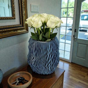 May include: A blue textured vase filled with white roses. The vase has a unique, wavy design and sits on a wooden surface. A small wooden bowl with keys is in the foreground. A mirror with a gold frame is visible in the background.
