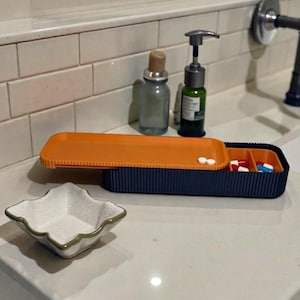 May include: A navy blue pill organizer with an orange lid, open to reveal pills in red, blue, and white. A small white ceramic dish and glass bottles are also on the counter.
