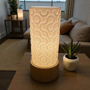 May include: A cylindrical table lamp with a cream-colored, textured shade and a beige base. The shade has a vertical, wavy pattern. A small potted plant sits to the right of the lamp on a wooden table. The lamp is lit, emitting a warm glow.
