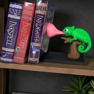 May include: A whimsical green chameleon bookend with a long pink tongue, appearing to lick a book. The bookend is perched on a brown tree branch and base, holding up three books with titles like "Inkspell" and "Inkheart".