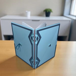 May include: A light blue notebook with a dark blue geometric border and a textured surface. The notebook is open, revealing white pages inside. It features a small blue elastic loop on the front cover and a central hinge mechanism with red accents.