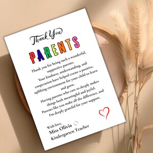 May include: A white card with the words "Thank You Parents" in colorful letters. The card includes a message of gratitude from a kindergarten teacher, with a red heart at the bottom. The card is surrounded by a wooden frame and dried plants.