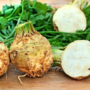 Könnte beinhalten: Nahaufnahme von frischen Knollensellerie und grünen Sellerieblättern auf einer Holzoberfläche. Der Knollensellerie ist hellbraun mit rauer Textur, und einer ist halbiert, wodurch das weiße Innere sichtbar wird.