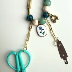 May include: A gold-toned keychain with a variety of charms. The keychain features a teal pair of scissors, a small heart charm, and a floral pendant. The chain also has a series of beads in shades of white, teal, and green.
